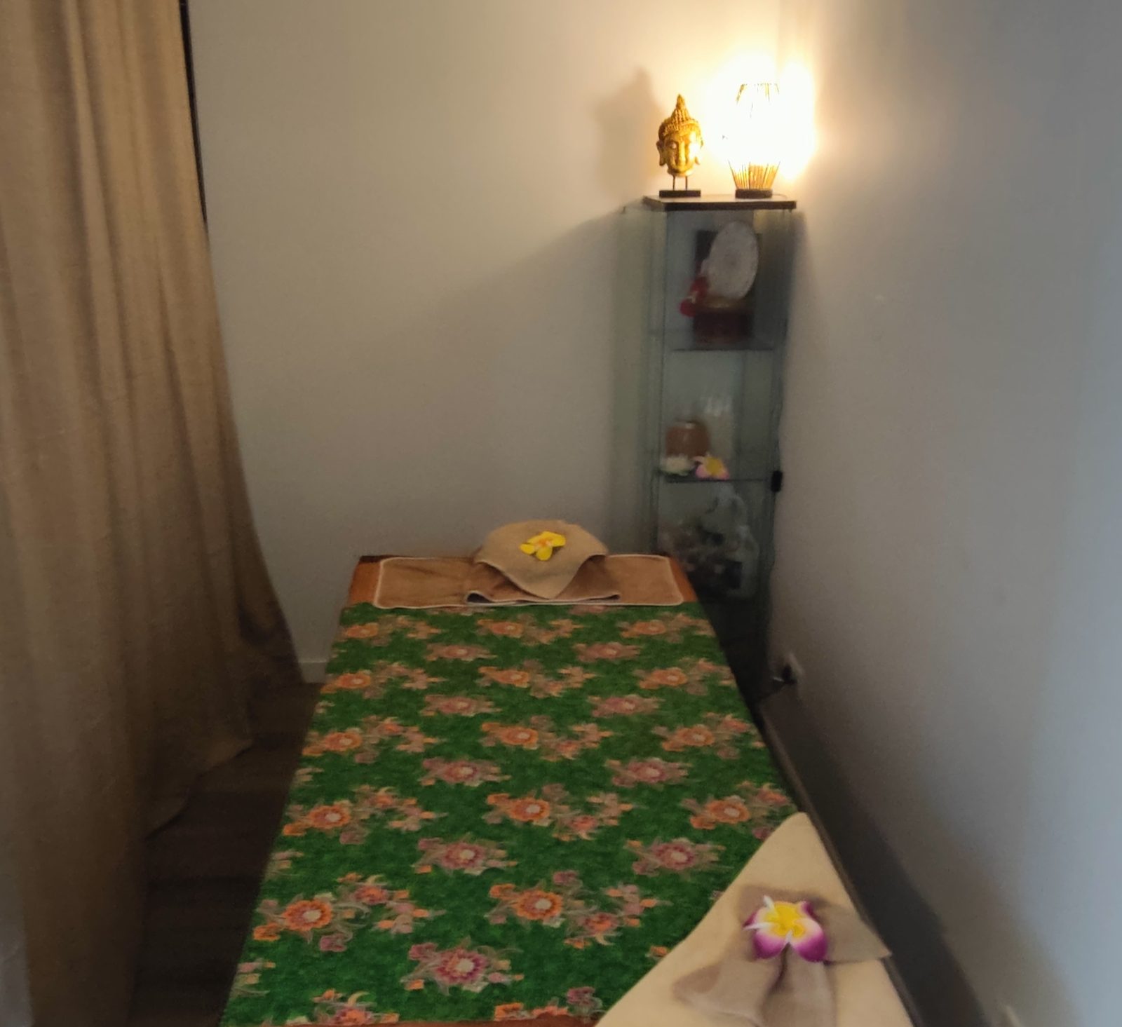 Een behandelruimte met een groene Thaise sprei met bloemen, zacht lamplicht en linnen gordijnen